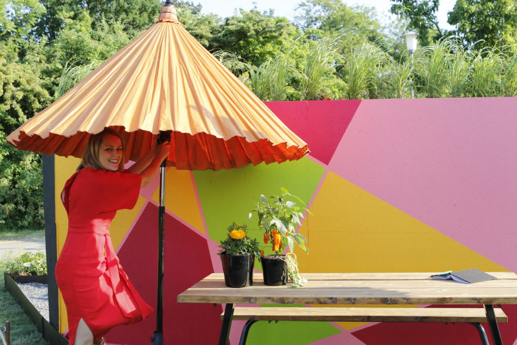 Cecilia Karlsson under parasoll vid ett picknickbord i utställningsträdgården på Malmö Garden Show 2018, framför en färgstark vägg och prydnadsgräs.