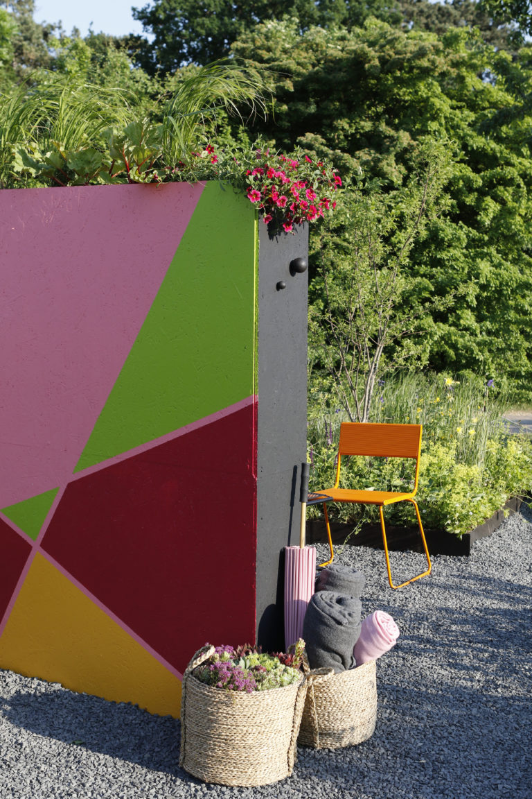 Färgstark planteringsvägg med prydnadsgräs och rosa blommor, grusgång och orange stol i utställningsträdgården på Malmö Garden Show 2018.