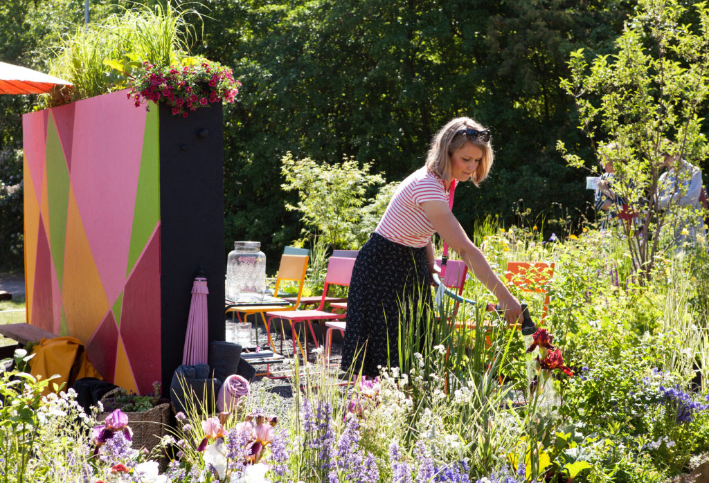 Färgstark utställningsträdgård på Malmö Garden Show med blommande perenner, målade väggar och sittplats med färgglada stolar.