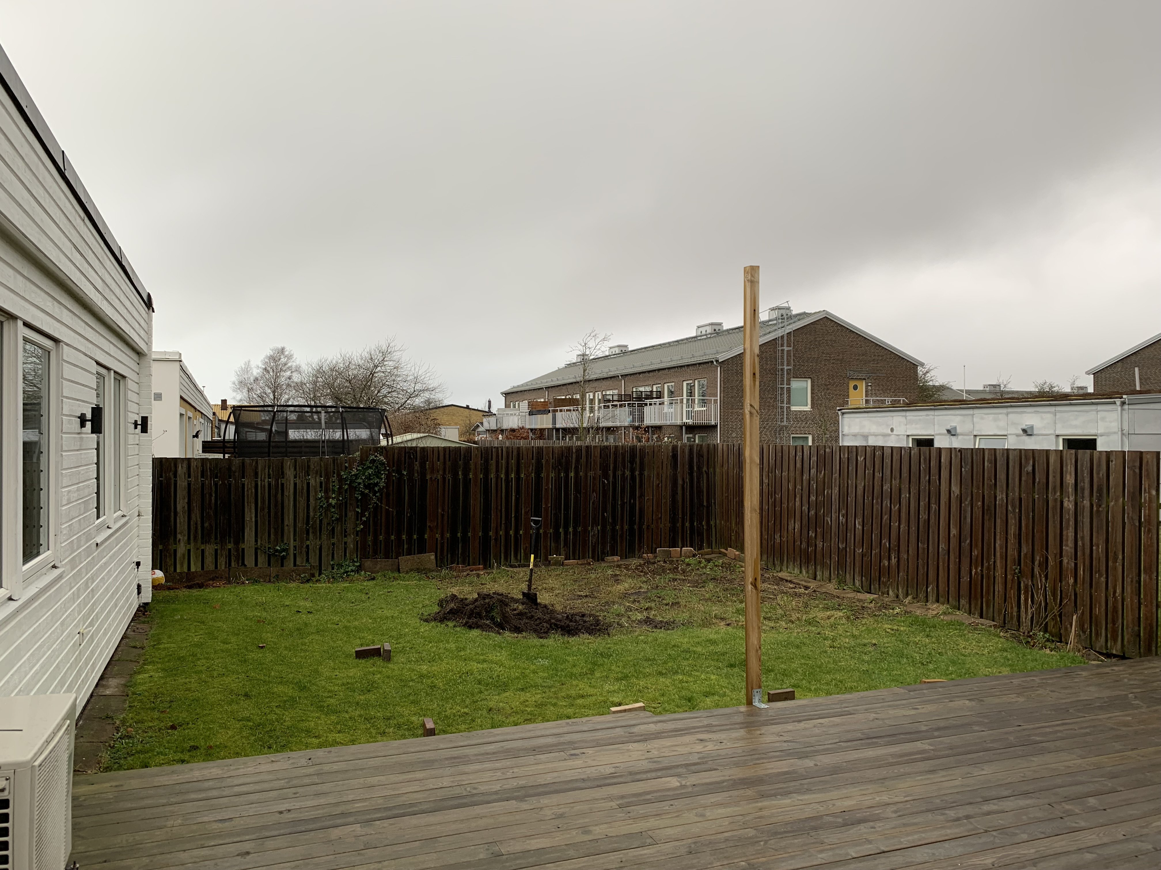 Bakgården före omgestaltning: liten gräsmatta, trätrall och högt trästaket, med några nygrävda planteringsytor.