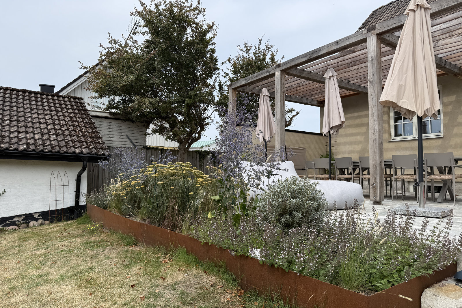 Frodig perennplantering i cortenstål ramar in uteplatsen med pergola, matbord och parasoller i villaträdgården i Mölle.